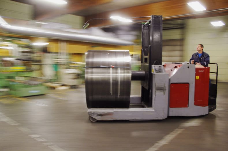 Operator driving a material handling forklift, moving a large metal coil through an industrial warehouse with motion blur.