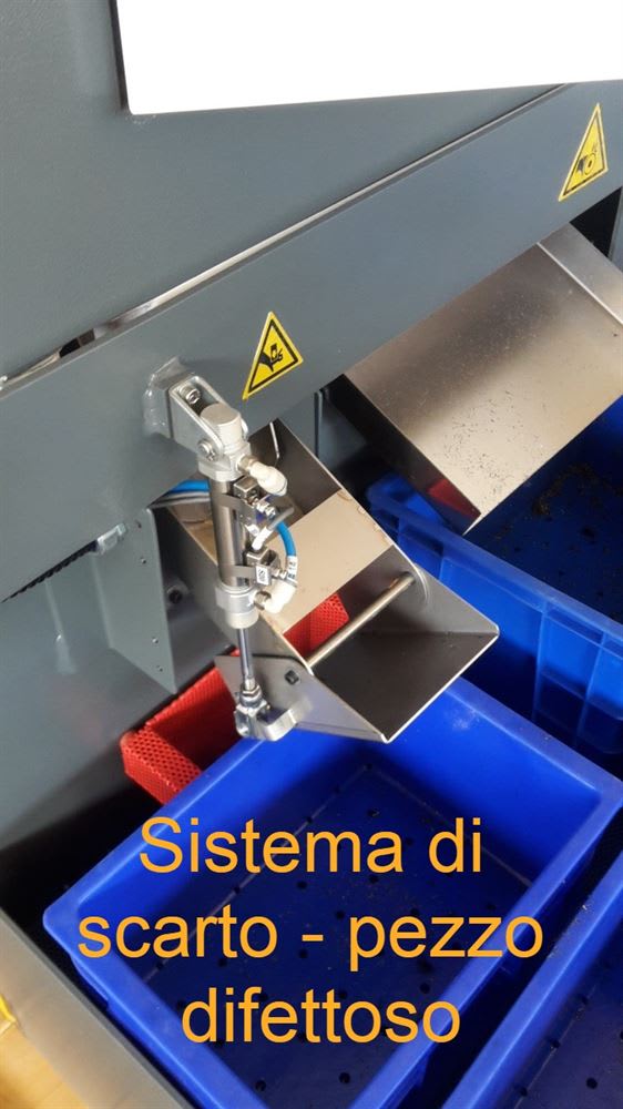 Sistema automatico di scarto per pezzi difettosi su macchinario industriale, con attuatore pneumatico che devia gli elementi in un contenitore di raccolta blu.