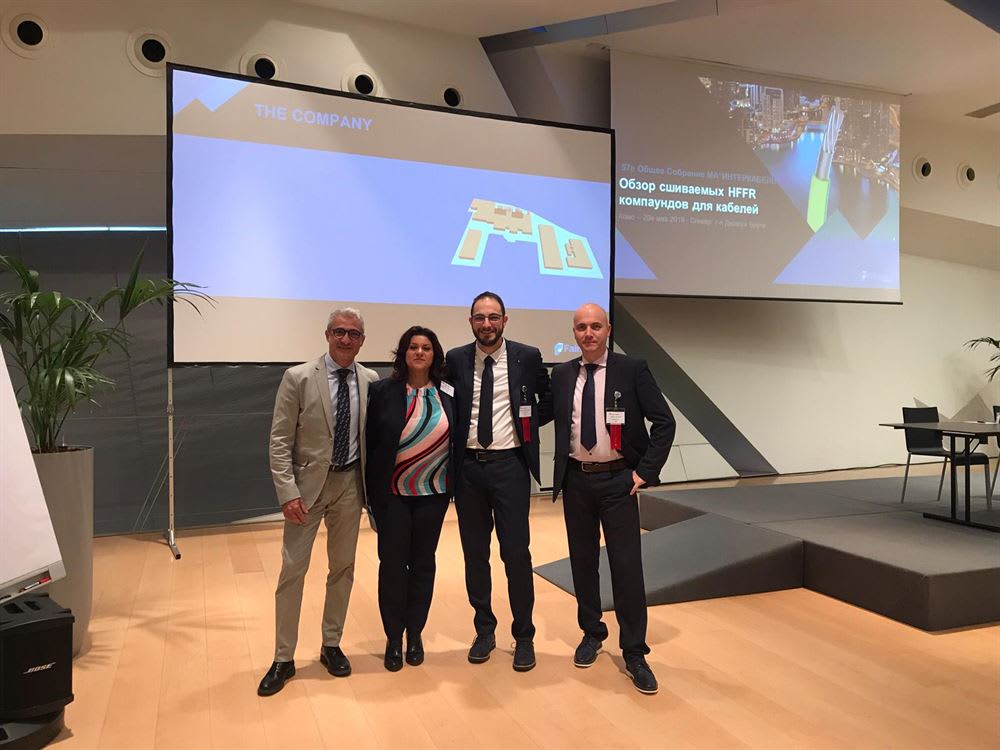 Gruppo di quattro professionisti sorridenti, tre uomini e una donna, in piedi davanti a schermi di proiezione durante una conferenza aziendale. Gli schermi mostrano presentazioni su 'THE COMPANY' e sui compound HFFR per cavi.