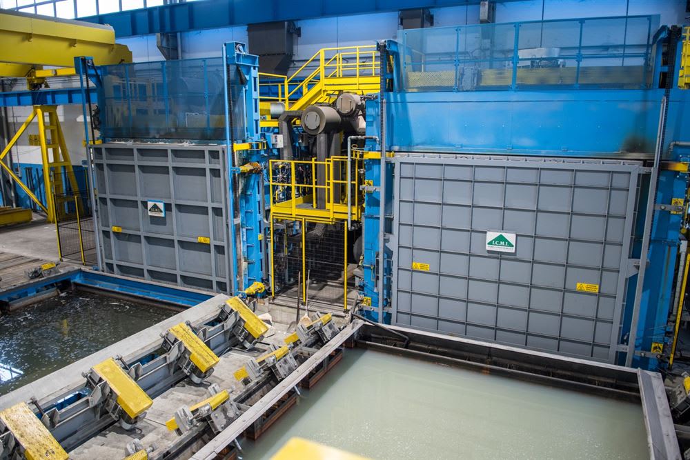 Large industrial plant interior with blue and yellow heavy machinery, two grey paneled processing units, and foreground tanks containing liquid for material treatment. The grey units display warning signs and an ICML logo.