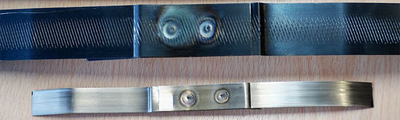 Two multi-segment metal strips: a dark blued strip with diagonal textured ridges and two heat-discolored spot welds, positioned above a lighter brushed strip with two mechanical joins.