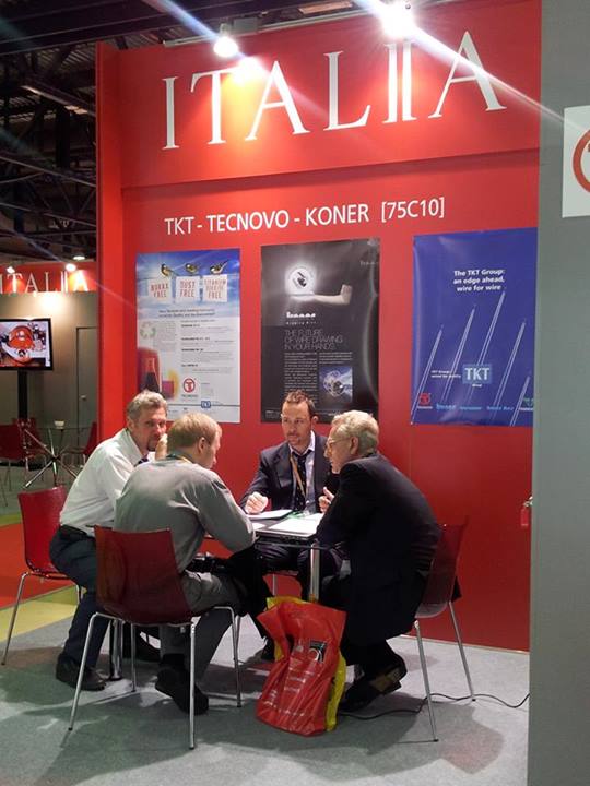 Incontro d'affari tra quattro uomini seduti a un tavolo presso lo stand fieristico rosso con il logo ITALIA TKT-TECNOVO-KONER.