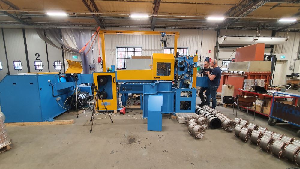 Large blue and yellow industrial wire processing machine in a factory, with two technicians monitoring operations and spools of finished wire on the floor.