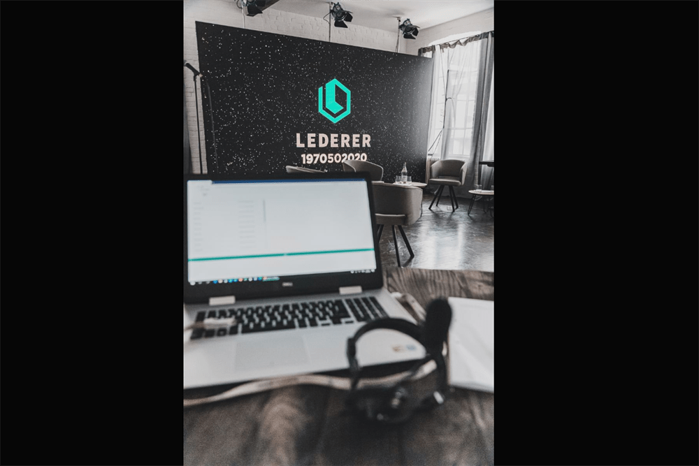 Laptop and headset in the foreground, out of focus, with a stage setup in the background. The stage features a black backdrop with a bright teal geometric logo, the word 'LEDERER', and '1970502020' text. Two chairs, a small table with a water bottle, and stage lights are visible, indicating a virtual event or presentation.