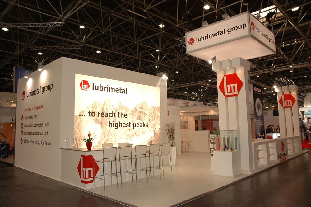 Современный белый выставочный стенд Lubrimetal Group с красными логотипами в выставочном зале. На заднем плане светящийся баннер с горным пейзажем и надписью «…to reach the highest peaks». Перед ним барная стойка со стульями. По бокам расположены высокие колонны с логотипом LM и подвесная вывеска «lubrimetal group».