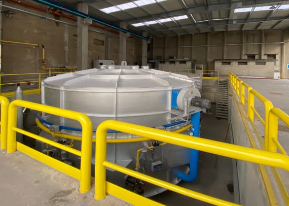 Grande serbatoio di processo industriale in metallo argentato con tubazioni blu e gialle, circondato da ringhiere di sicurezza gialle, all'interno di un capannone industriale moderno.