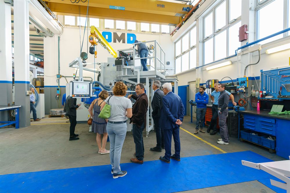 A group of professionals, including visitors and staff, observing a large OMD industrial machine in a bright, modern factory workshop, with a technician on the machine platform and workbenches with tools visible.