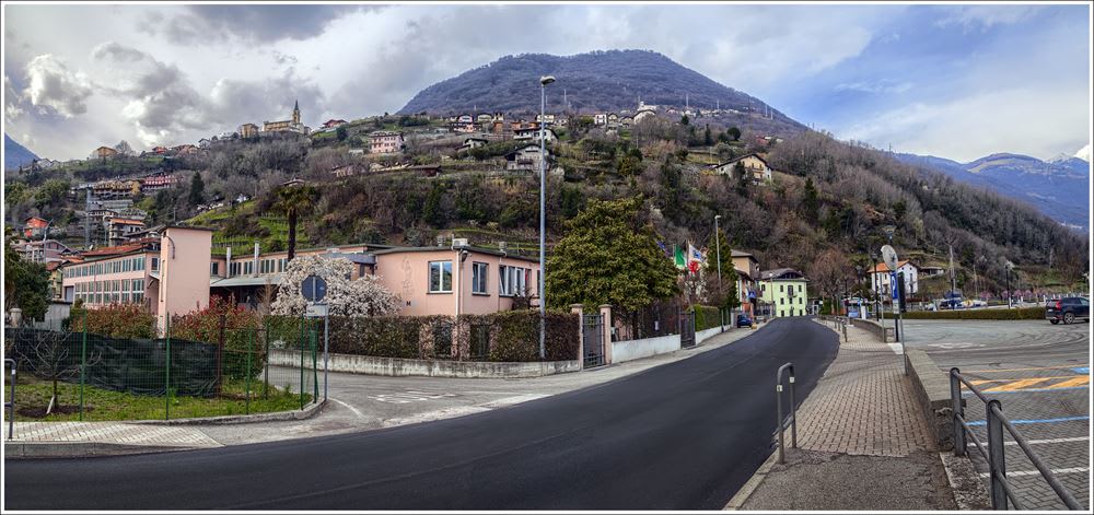 Panorama di un villaggio montano italiano. In primo piano, una strada asfaltata e diversi edifici, tra cui un grande complesso rosa e una casa verde chiaro, con bandiere italiane visibili. Il villaggio si estende su una ripida collina alberata, con numerose abitazioni sparse. In cima alla collina spicca una chiesa con campanile. Sullo sfondo, montagne maestose si stagliano sotto un cielo parzialmente nuvoloso.