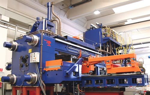 Large blue and orange heavy industrial machine featuring a prominent TP logo, complex hydraulic systems with pipes, and worker access platforms with yellow railings, set within a manufacturing facility.