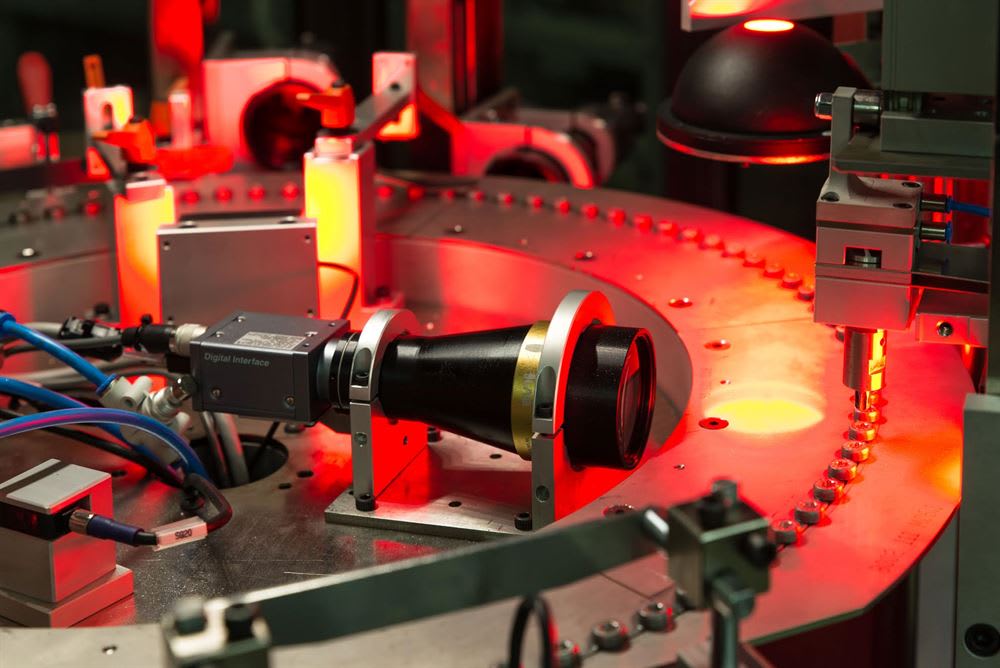 Optical inspection systems: a light gray OptoGaugeLine NANO 5AI by Regginspection inspecting a bolt, and a dark gray machine inspecting a pin, both showing detailed precision measurement data on monitors.