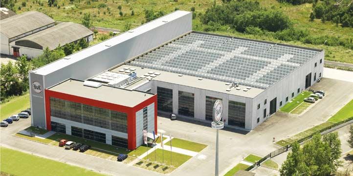 Veduta aerea del complesso industriale Rivit S.p.A. con ampio tetto a pannelli solari che forma il logo aziendale e una palazzina uffici moderna dalla facciata rossa. Parcheggi e aree verdi circondano lo stabilimento.