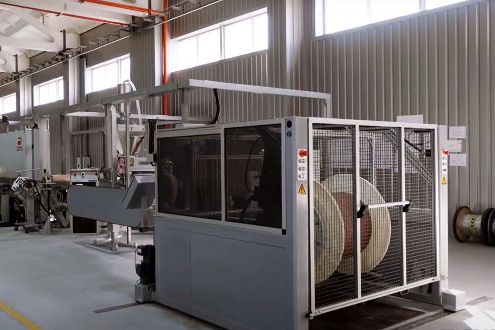Industrial manufacturing facility with a production line machine and large wire spools inside, featuring high windows and corrugated metal walls.