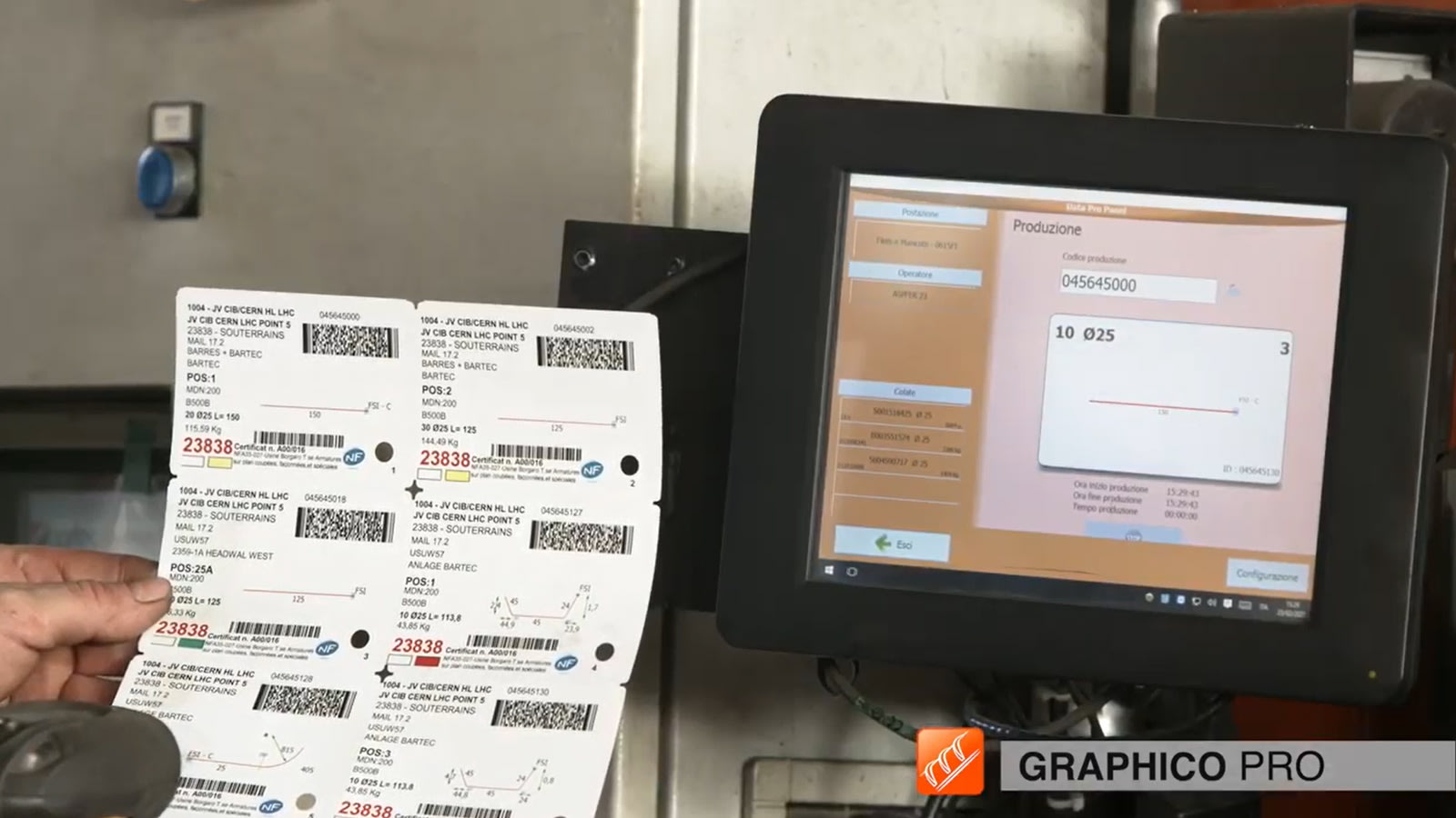 Mano sosteniendo una hoja de etiquetas con códigos de barras para componentes del CERN LHC, frente a una pantalla táctil industrial que muestra datos de seguimiento de producción y temperatura.