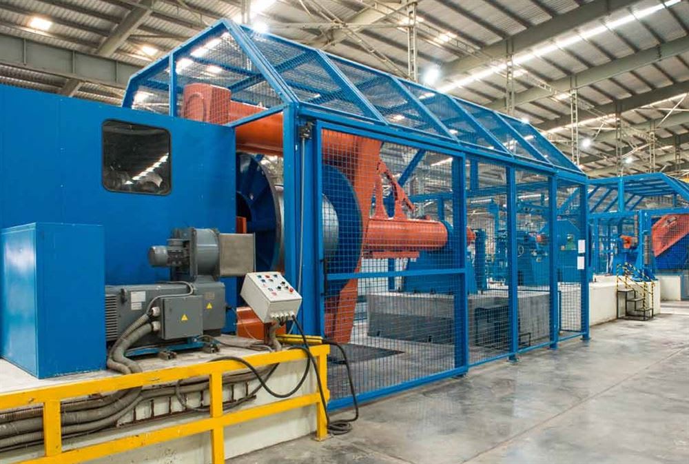 Blue and orange industrial machinery for cable or wire manufacturing, featuring large rotating components and mesh safety guards, inside a brightly lit factory.