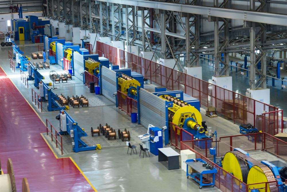 Modern industrial factory with a cable and wire manufacturing production line, featuring large blue and yellow winding machines, safety fences, and spools of copper wire on the polished floor.