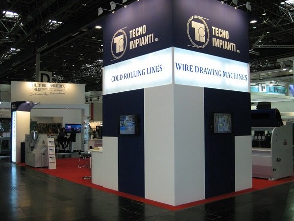 Stand d'exposition TECNO IMPIANTI SRL à un salon professionnel, affichant des machines de laminage à froid et de tréfilage avec des écrans vidéo.