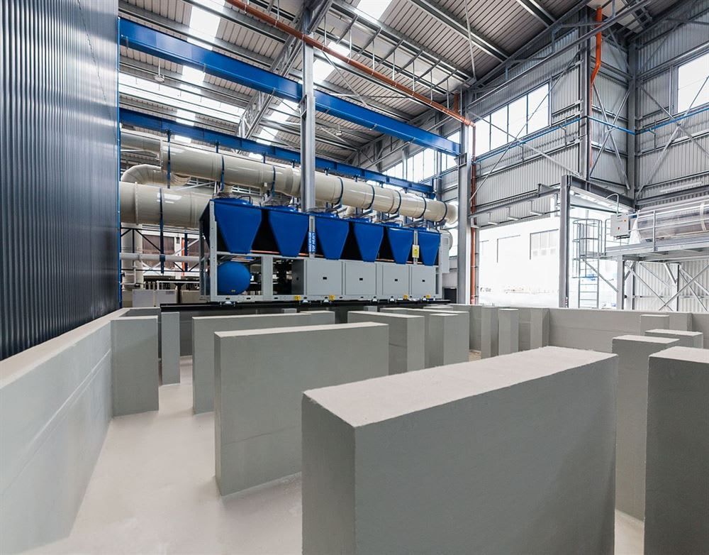 Interior of a modern industrial plant featuring large blue industrial cooling units connected by white pipes, with a foreground of grey concrete partitions arranged in a structured layout.