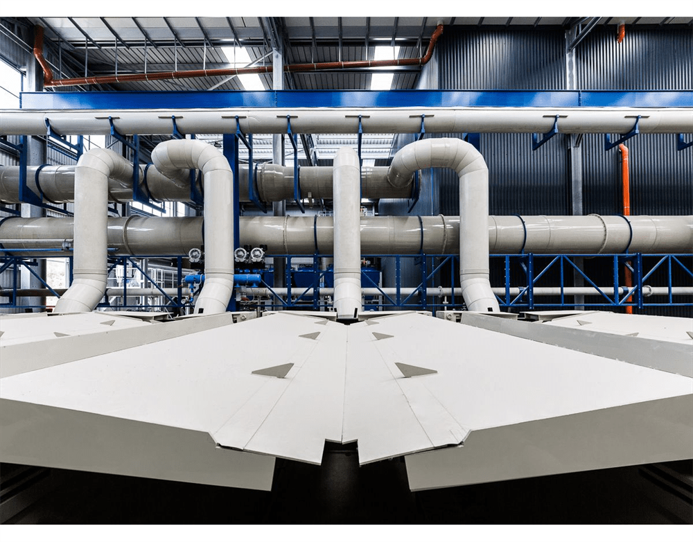 Modern industrial facility interior featuring prominent white processing pipes, blue structural supports, and large angular white machinery in the foreground.