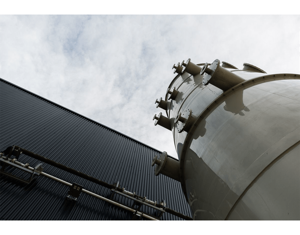 Industrial processing facility featuring a large, light-colored cylindrical vessel with multiple pipe connections, standing against a dark corrugated metal building under a cloudy sky.