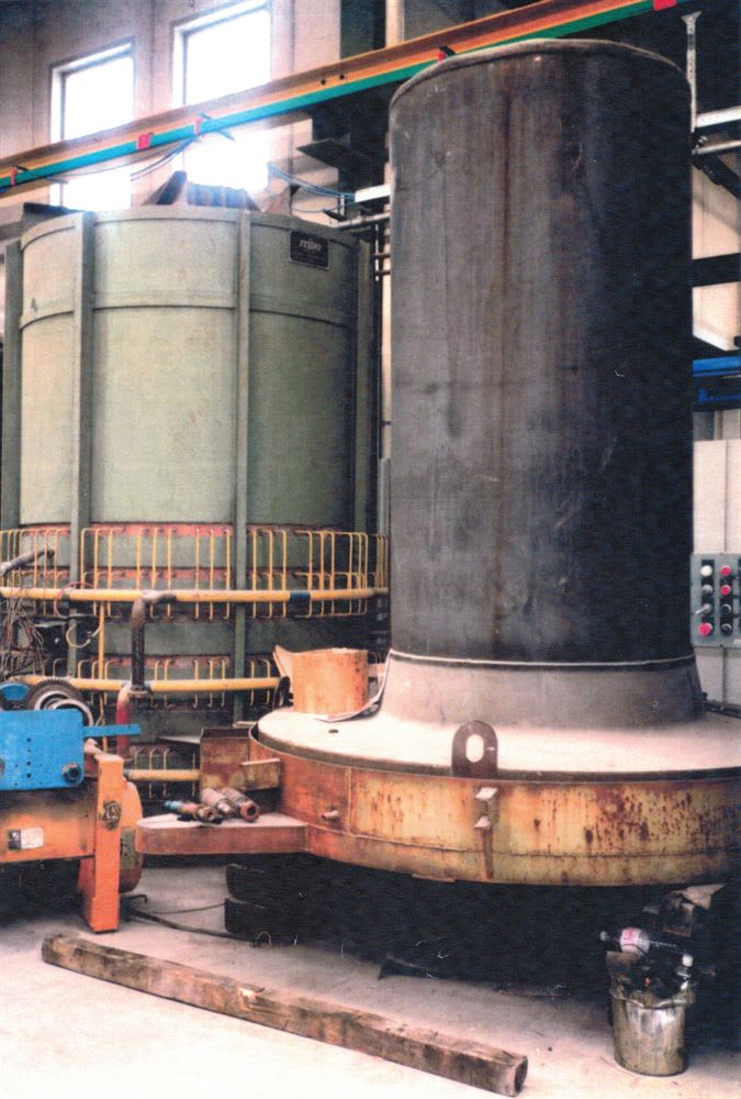 Un impianto industriale presenta due grandi strutture cilindriche. A destra, un imponente serbatoio metallico scuro con segni di usura e una base ampia e arrugginita. A sinistra, una serie di serbatoi più chiari con fasce di rinforzo e una ringhiera di protezione gialla intorno alla base. Si intravedono travi strutturali, tubazioni e un pannello di controllo su una parete.