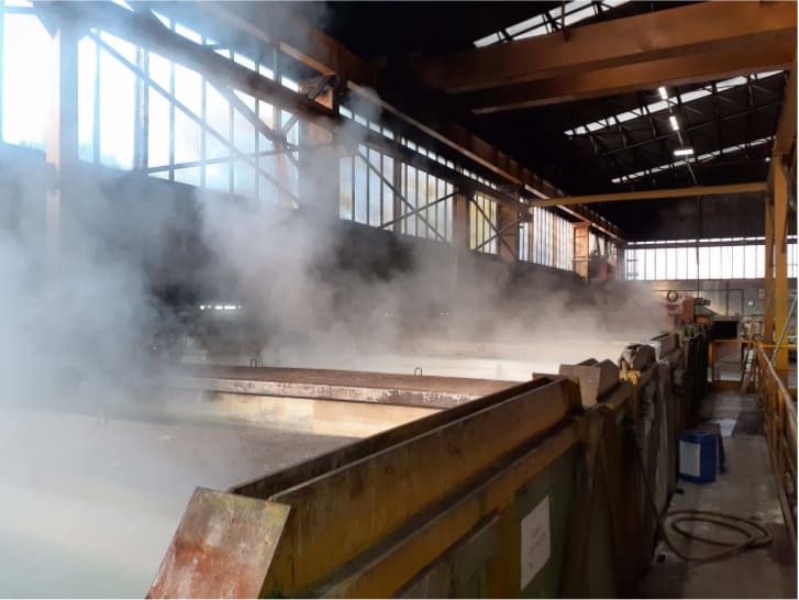 Interno di un capannone industriale con abbondante vapore che si innalza da grandi vasche di lavorazione, sotto un tetto con strutture di carroponte.