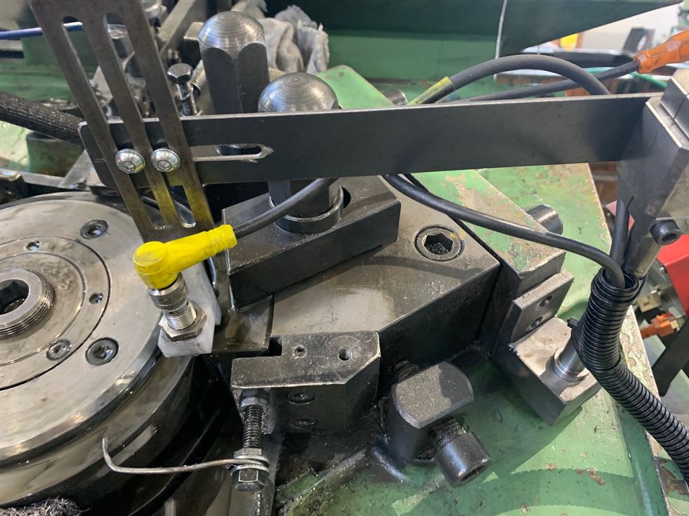 Dettaglio di macchinario industriale con sensore di prossimità giallo, cavi e componenti metallici per lavorazione di precisione.