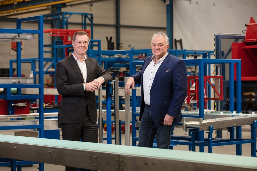 Two smiling business executives posing in a modern manufacturing facility with industrial automation and a conveyor belt in the foreground.