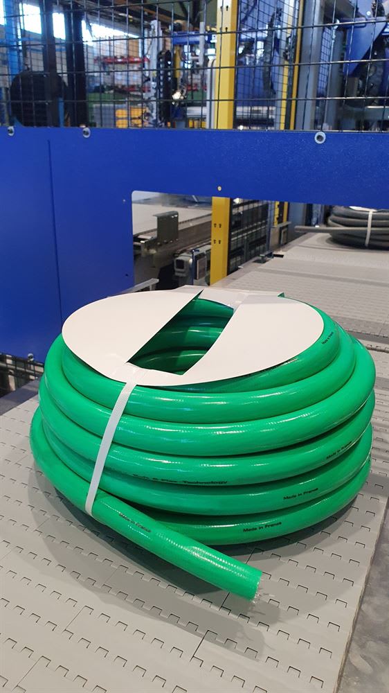 Bright green garden hose coil with 'Made in France' visible on its surface, secured by a white strap and label, resting on a grey tiled floor in an industrial manufacturing facility.