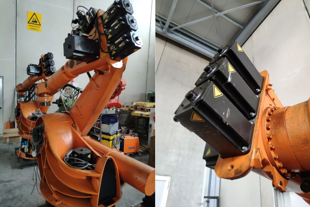 Robot industriale arancione con braccio articolato, dotato di un attrezzo finale composto da quattro moduli neri con simboli di pericolo gialli, situato in un ambiente di fabbrica con pareti chiare e segnaletica di 'Passaggio Carrelli'.