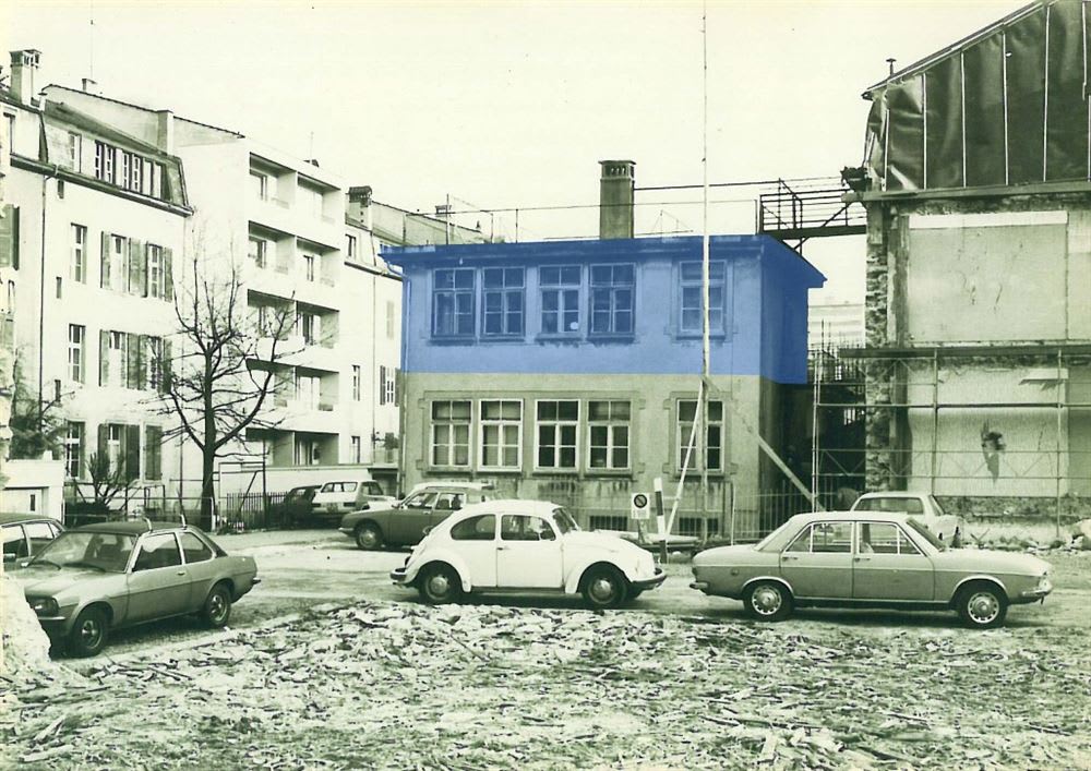 Urban street scene from the 1970s or 1980s featuring a two-story building flanked by a modern apartment block and a building undergoing demolition. Several vintage cars, including a white VW Beetle, are parked on a rough, unpaved lot in the foreground.