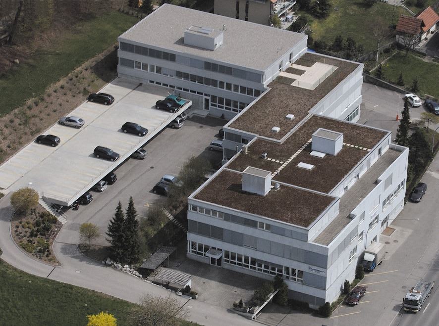 Aerial view of a modern multi-building corporate complex, featuring an elevated concrete parking structure with cars, and several buildings with flat, vegetated green roofs, surrounded by access roads and additional parking areas.
