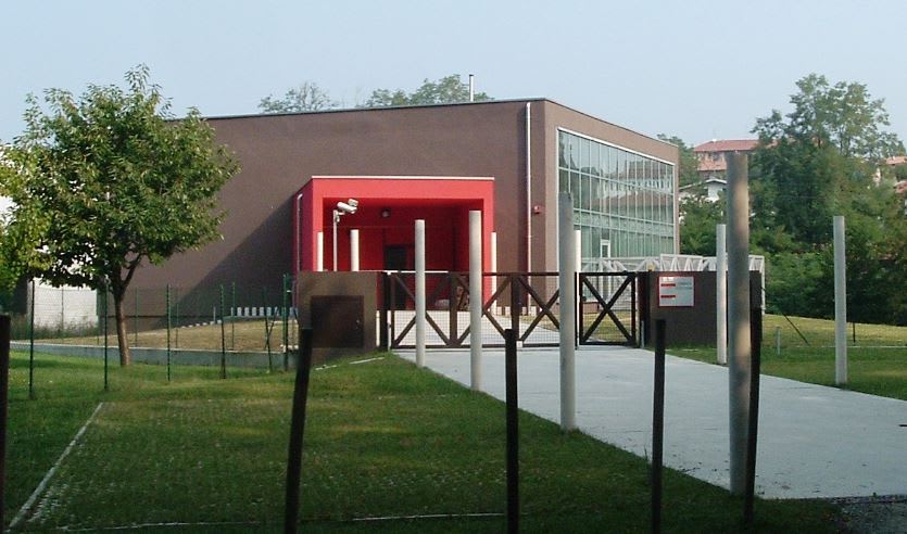 Modern brown building with a prominent red entrance and large glass windows, set amidst a green lawn, paved path, and several white fence posts.