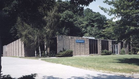 Modern commercial building with a textured stone facade, dark vertical windows, and a blue 'Sunhaven' sign, surrounded by a green lawn and tall trees under a clear sky.