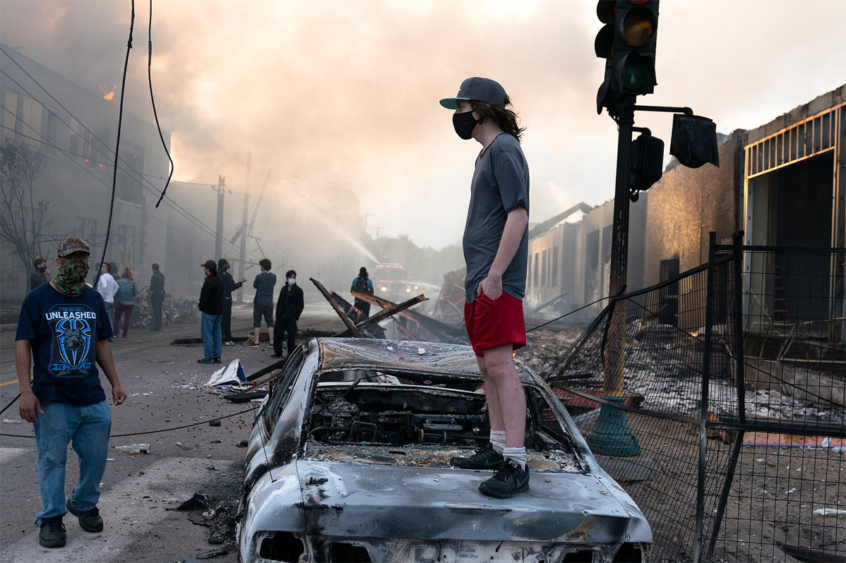 A man stands on a burned out car. Fires seen burning behind him at the Lake St area of Minneapolis, Minnesota.