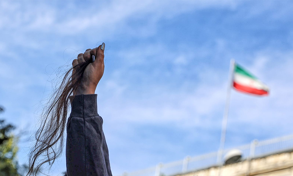 A photo of a woman's hand holding her hair after cutting it, with the Iranian flag in the background