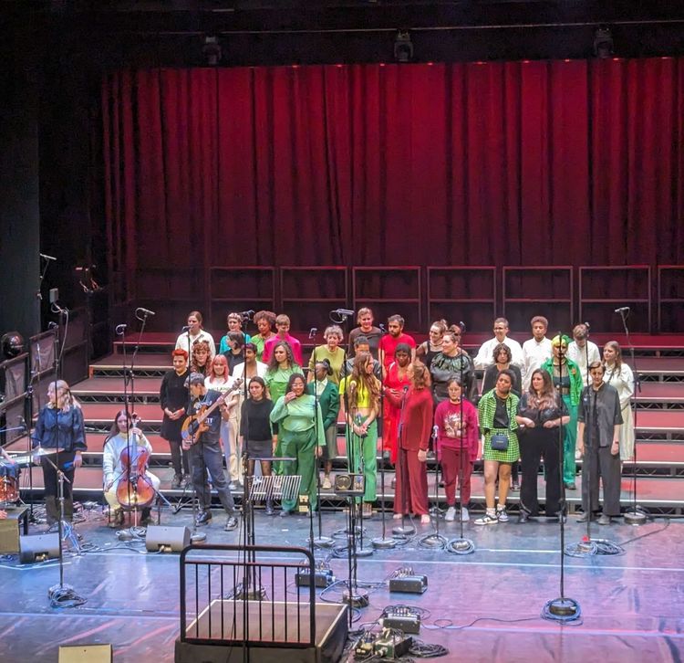 F*Choir on stage at the epic Hackney Emprie, as seen from the top of the theatre balcony. Singers are dressed in red, yellow, black, and green and preparing to sing for a crowd of 800 people