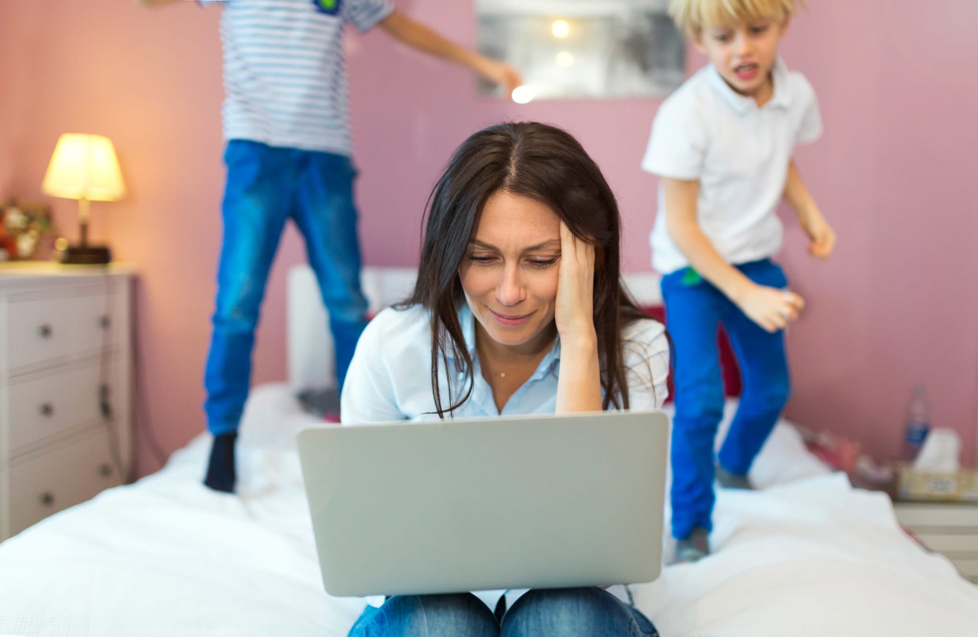 The-parents-are-not-okay Mother working from home on a laptop distressed while her two boys jump on the bed behind her