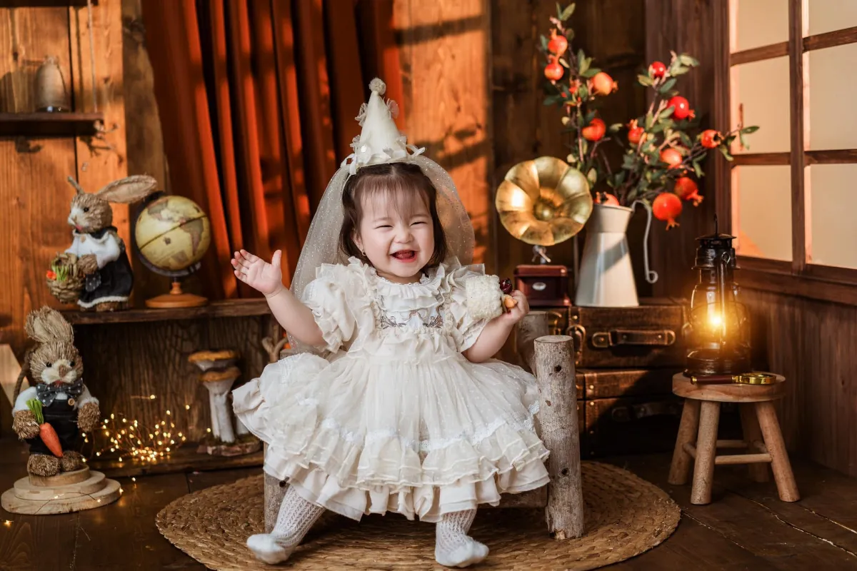 Bé ngồi chơi trên sàn gỗ trong váy trắng — sau tiêm, ba mẹ cần theo dõi sốt và phản ứng tại chỗ tiêm của bé