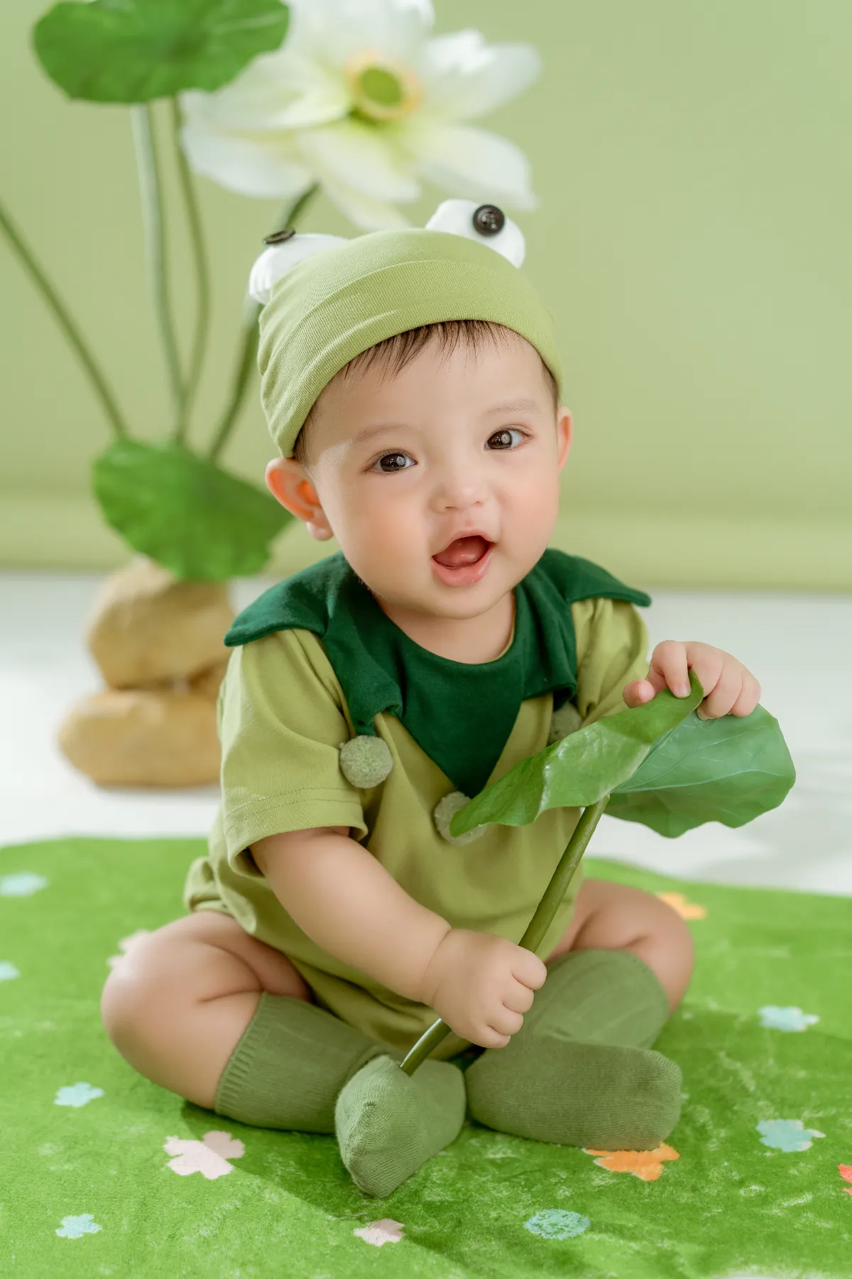 Bé ngồi chơi trong bộ đồ xanh lá — trước ngày tiêm, ba mẹ nên kiểm tra sức khỏe và sổ tiêm chủng của bé
