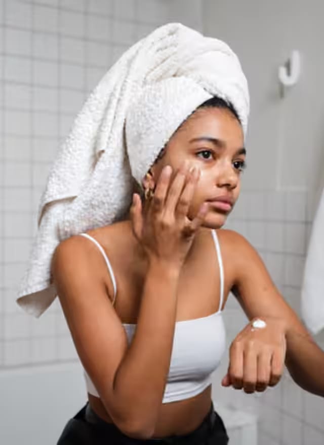 A woman applying cream to her cheek, with her full towel-wrapped head visible.