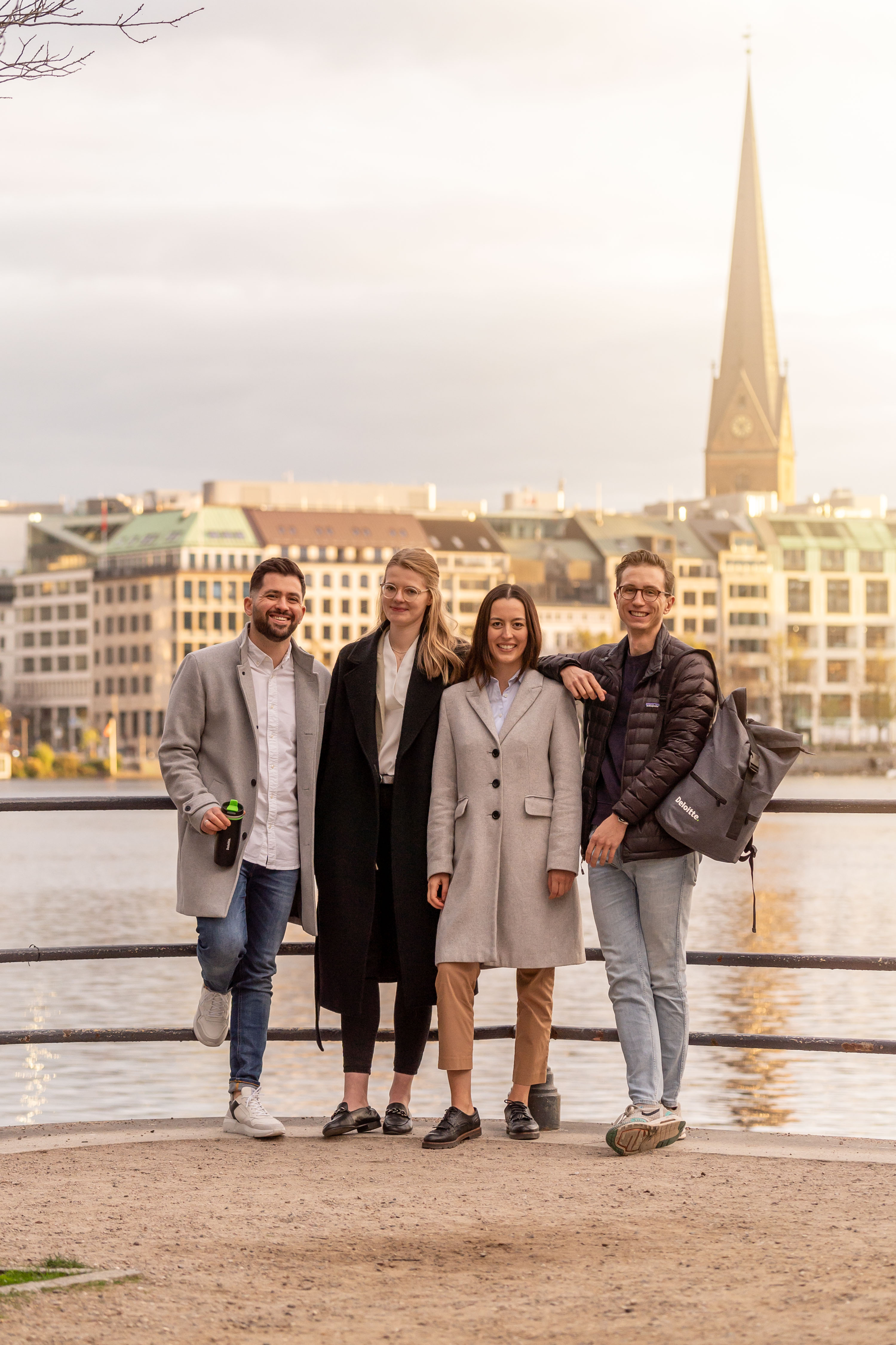 ier Personen stehen gemeinsam an einem Flussufer, im Hintergrund eine Stadtansicht mit Kirchturm.