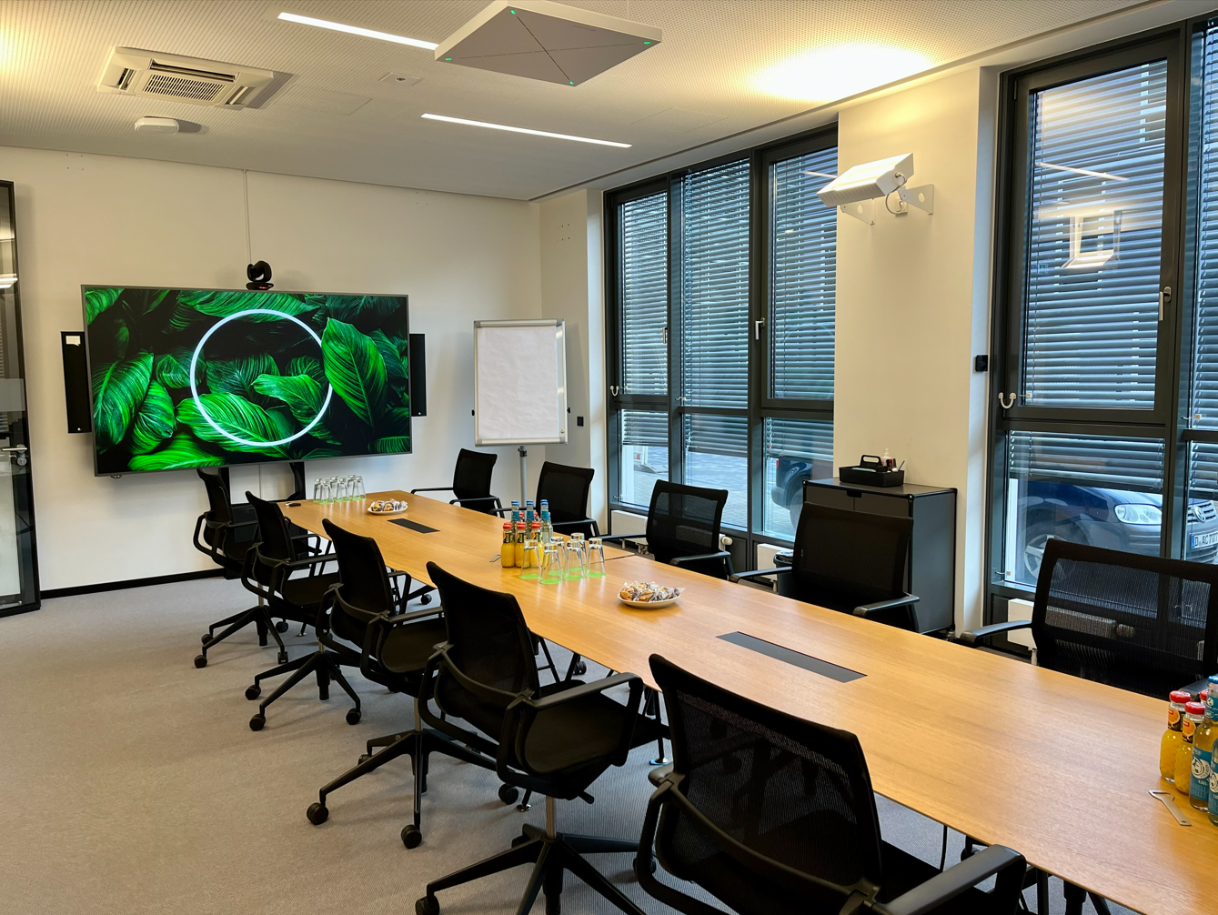 Moderner Konferenzraum mit großem Tisch, Bürostühlen, Bildschirm und Flipchart vor Fensterfront.