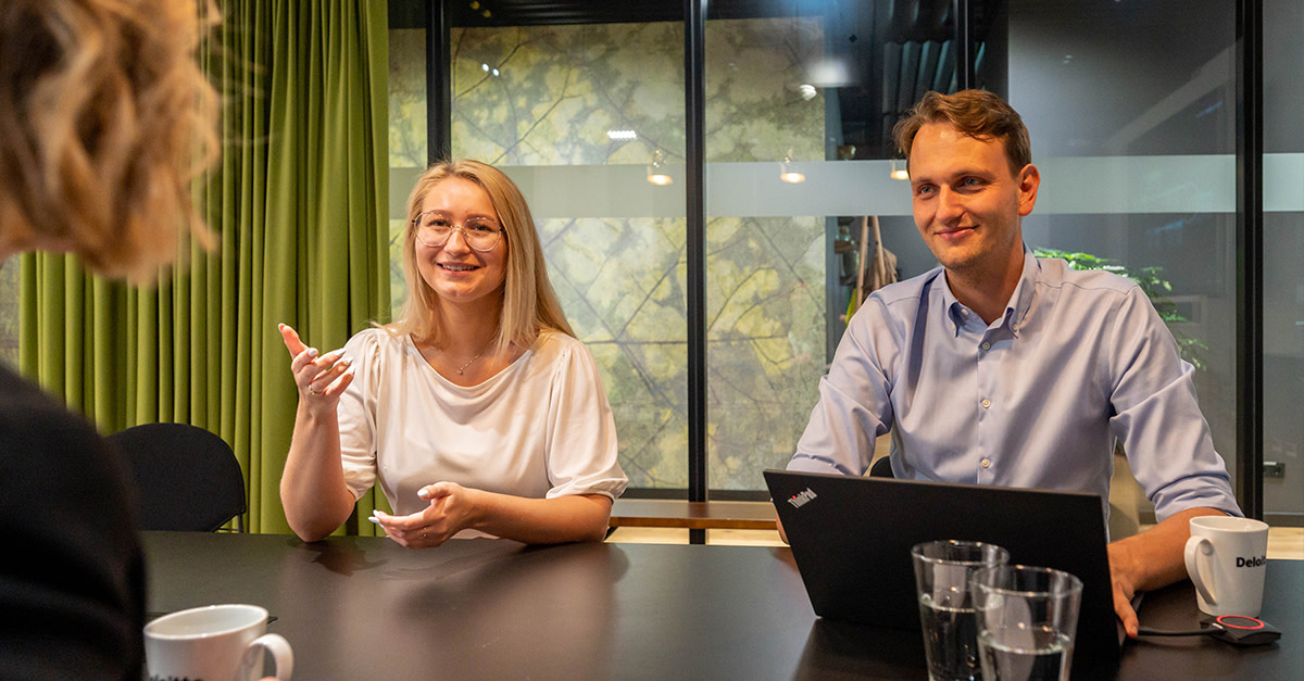 Drei Kolleg:innen führen ein Meeting am Tisch, mit Laptop und Getränken in moderner Umgebung.