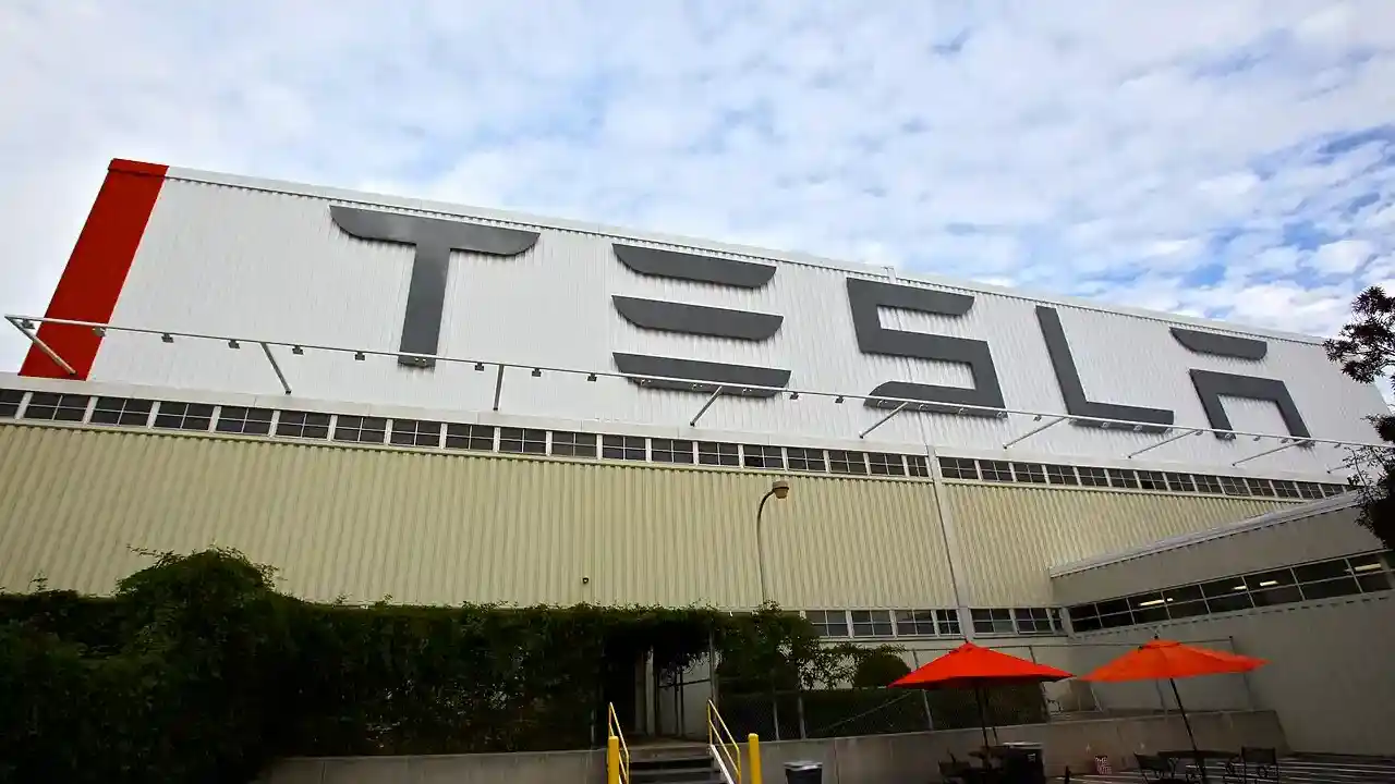 Tesla factory building with prominent logo on facade, surrounded by greenery and orange umbrellas, captured in 2025.