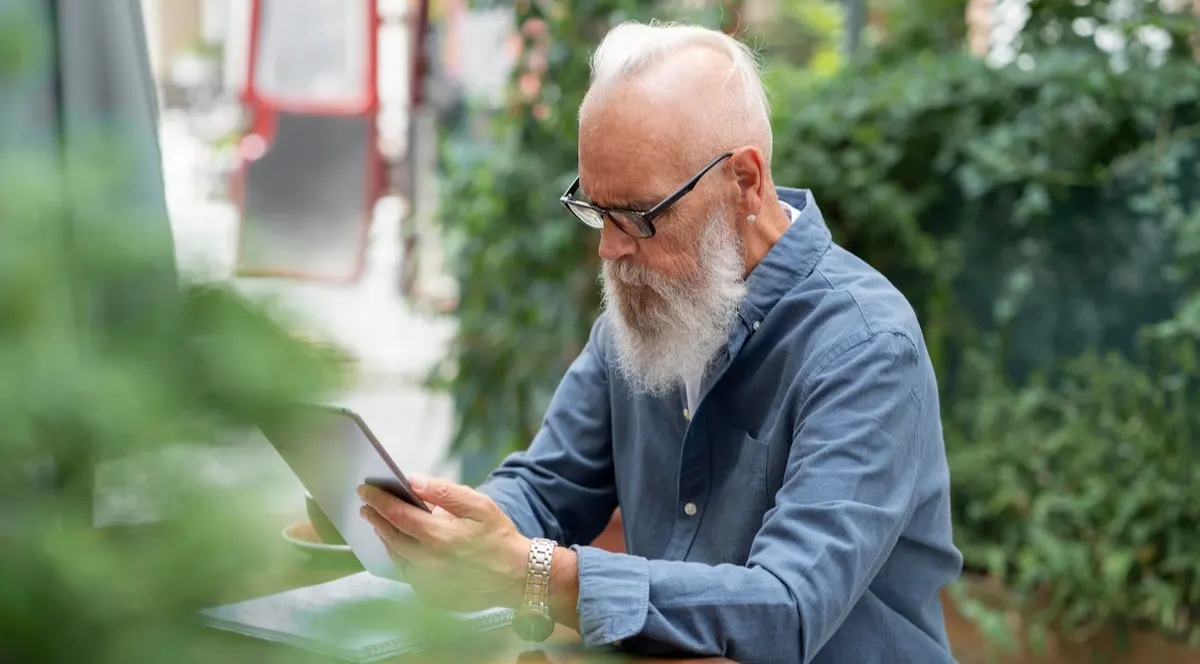 Senior man with beard and glasses using a tablet outdoors at a café table