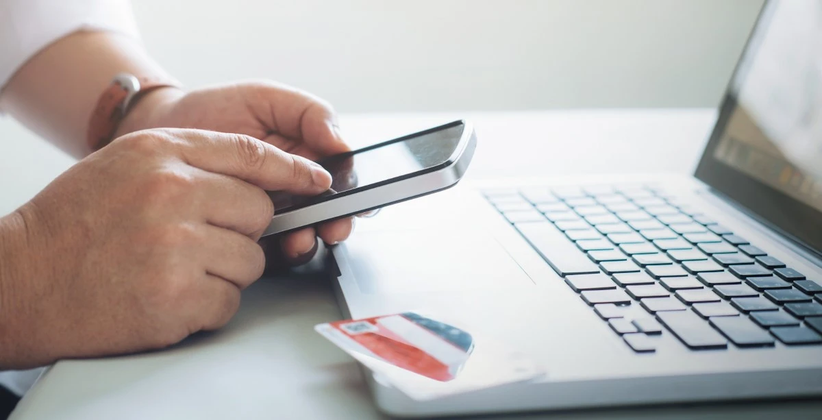 Hands holding a smartphone in front of a laptop with a debit card nearby, showing mobile banking activity