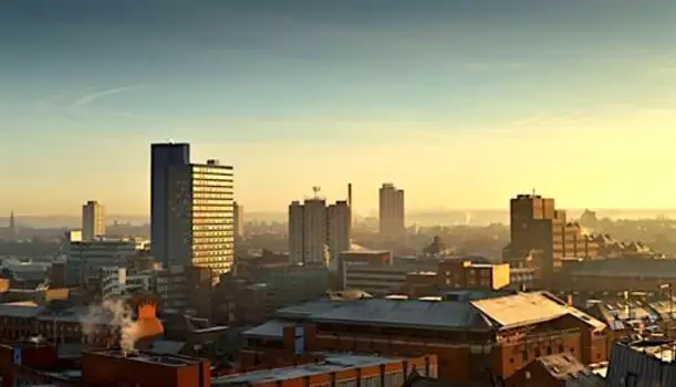 Skyline of Leicester city at sunrise, featuring a mix of modern high-rise buildings and older brick structures under a clear sky with a warm golden glow.