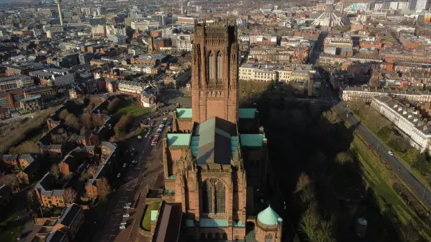 Aerial view of Liverpool Cathedral, a large sandstone structure with greenish copper roofs, standing prominently in the foreground. The cathedral is surrounded by residential streets, green spaces, and a dense urban cityscape. Liverpool landmarks such as 