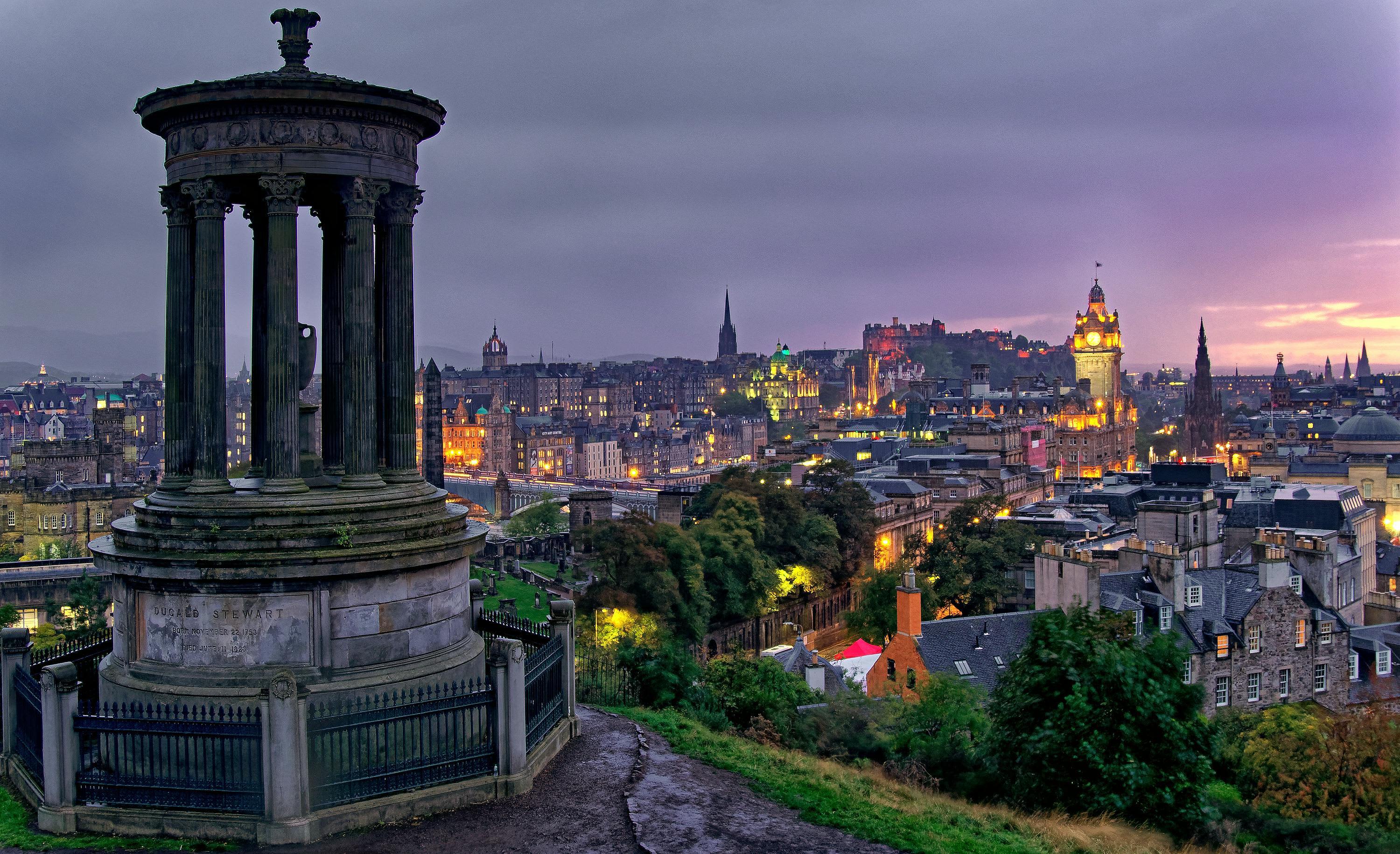 View of Edinburgh at dusk from Calton Hill, featuring the Dugald Stewart Monument in the foreground and the city&rsquo;s historic skyline, including Edinburgh Castle and the Balmoral Hotel clock tower under a dramatic purple sky.