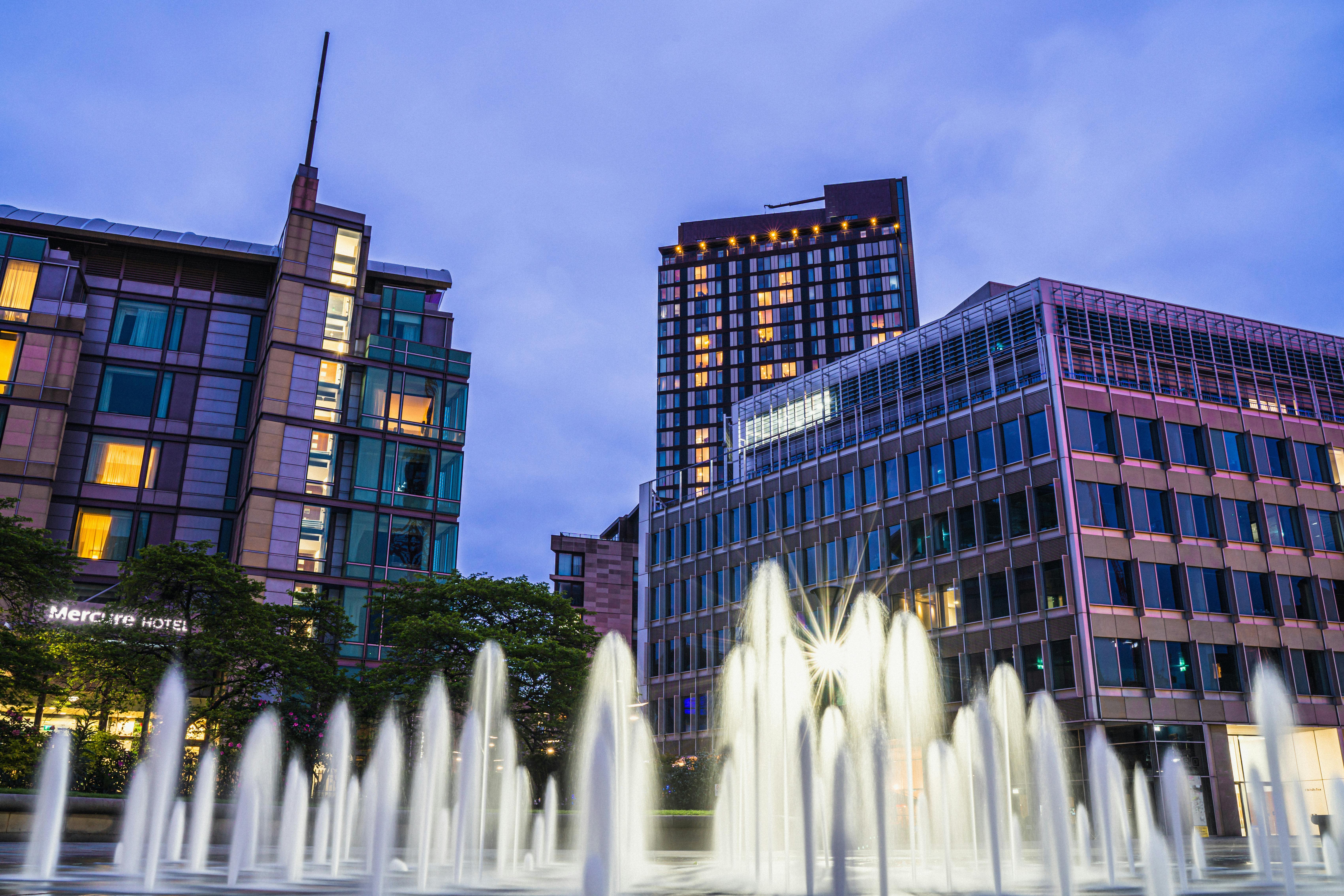 Fountain in Sheffield city centre at dusk, with modern glass buildings and the Mercure Hotel illuminated against a blue evening sky.